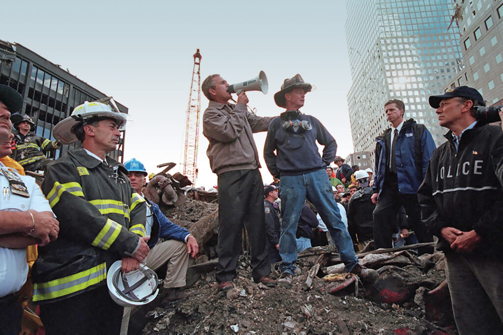 President George W. Bush stands on a pile of rubble and speaks through a microphone. Surrounding him are rescue workers and police. 